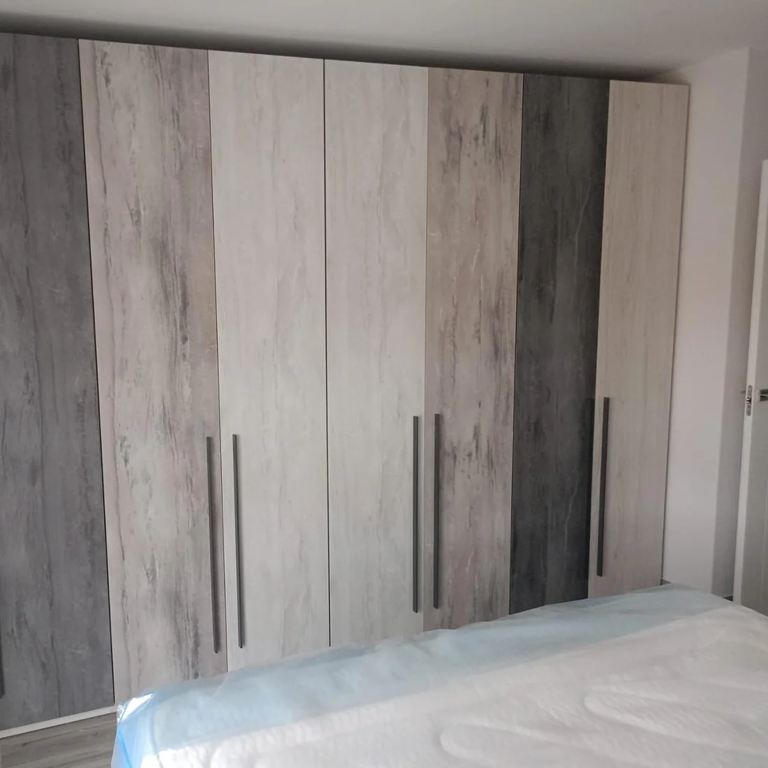 Frente de armarios a medida en un dormitorio, con puertas hasta el techo que alternan acabados en madera clara y piedra gris.