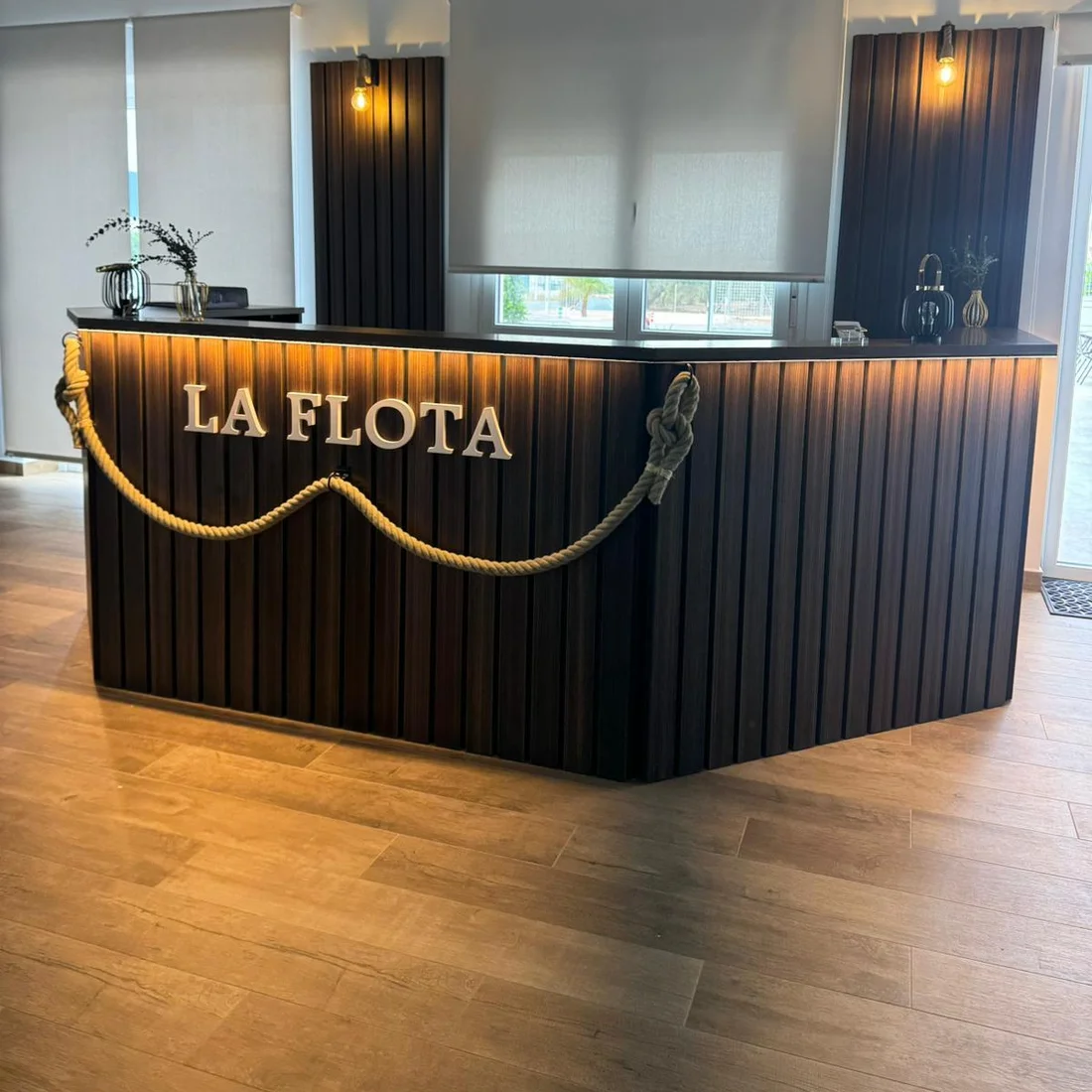 Mostrador de recepción a medida en madera oscura con frente de listones, letras retroiluminadas y una maroma cruzando el frontal.